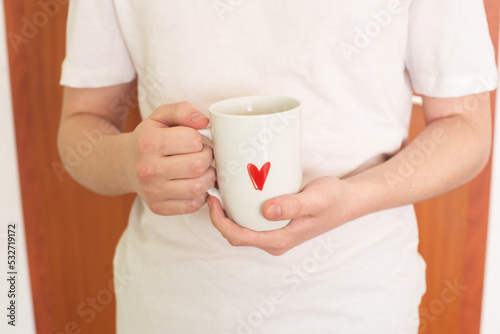 The hands of a young man in a white T-shirt hold a white cup with a red heart pattern. Tea time. Coffee time. Life style