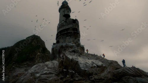 POV from tourist boat approach Cape Aniva, where located abandoned lighthouse, tourists climb rope to lighthouse, seagulls soar in air, Sakhalin Island photo