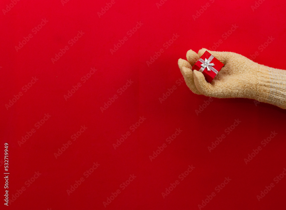 Squeezing a small red box with a Christmas present in her palm, a woman's hand in beige mittens looks beautiful on a red background. Concept for Christmas and New Year cards and backgrounds. Squeezing a small red box with a Christmas present in her palm, a woman's hand in beige mittens looks beautiful on a red background. Concept for Christmas and New Year cards and backgrounds.