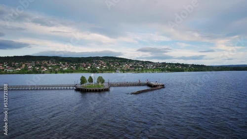 Aerial orbit shot. A pier at Lake Siljan in Sweden, people walk on the promenade. View of the town of Rättvik against the mountain and a harbor with sailing boats. Cloudy during sunset photo