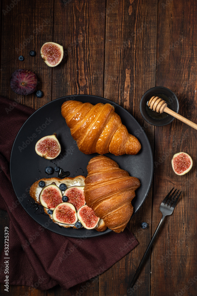 Croissants with cream cheese, figs, blueberries and honey on a black plate. Breakfast concept, top view.