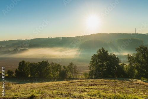 A view over a valley in the rolling hill landscape outside Maastricht, where the valley is covered with layers of fog and creating a nice setting with the sun beams through the mist over the fields