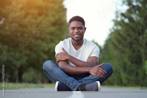 African American man sitting cross legged on asphalt shows toothy smile and enjoys life. Young guy sits against blurry trees in park at twilight © lenblr