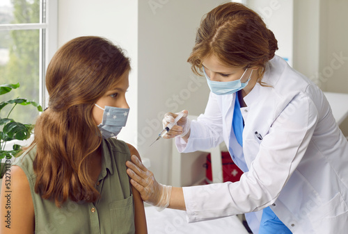 Doctor giving shot to child in face mask. Female nurse in white coat and facemask holding syringe and giving vaccine injection to girl during mandatory mass vaccination immunization campaign at school