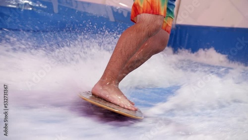 Attractive Man riding surf board on wave simulator indoors. Young surfer during training on generated waves. Water sport activity. Surfer guy enjoy surfing on indoor surf imitator.Close-up
