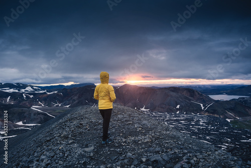 Hiker woman in yellow jacket standing on volcanic mountain and midnight sun from Blahnjukur trail in Icelandic highlands