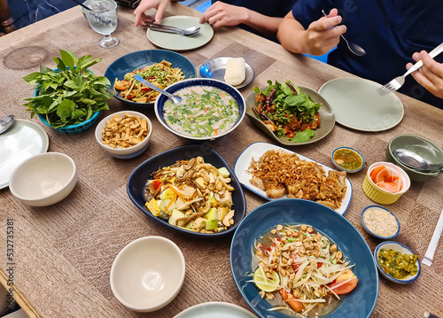 Variety of Thai food with papaya spicy salad, fruit salad, crispy pork, spicy pork soup, sticky rice and cutlery on wooden table