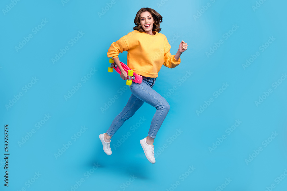 Full length photo of sweet cute girl dressed yellow pullover holding skateboard jumping high isolated blue color background