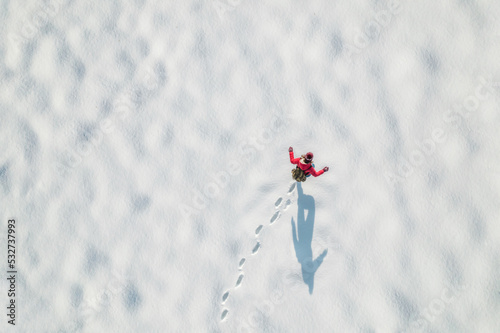 Woman in a red christmas jacket with backpack walking on the snow and doing footprints in a park. Winter. Drone, top, aerial view.