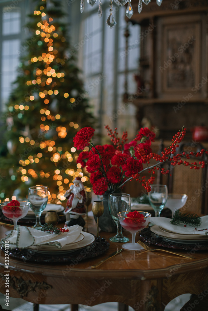 Still life of aesthetic table setting with red Christmas decorations. Festive holiday season. Christmas tree on background.
