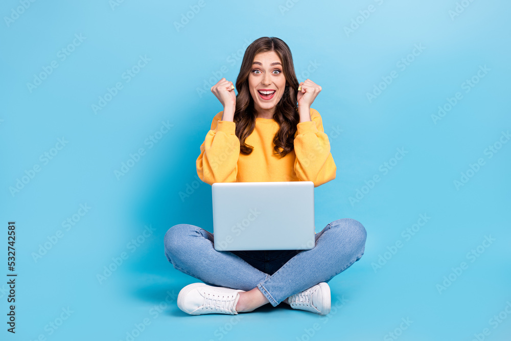 Full length photo of impressed lucky lady wear yellow sweater working modern device sitting legs crossed isolated blue color background Full length photo of impressed lucky lady wear yellow sweater working modern device sitting legs crossed isolated blue color background