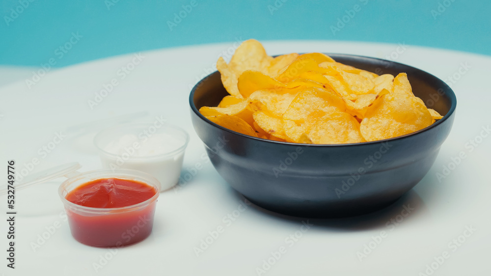 crunchy and ridged potato chips in bowl near plastic containers with ketchup and mayonnaise on blue.