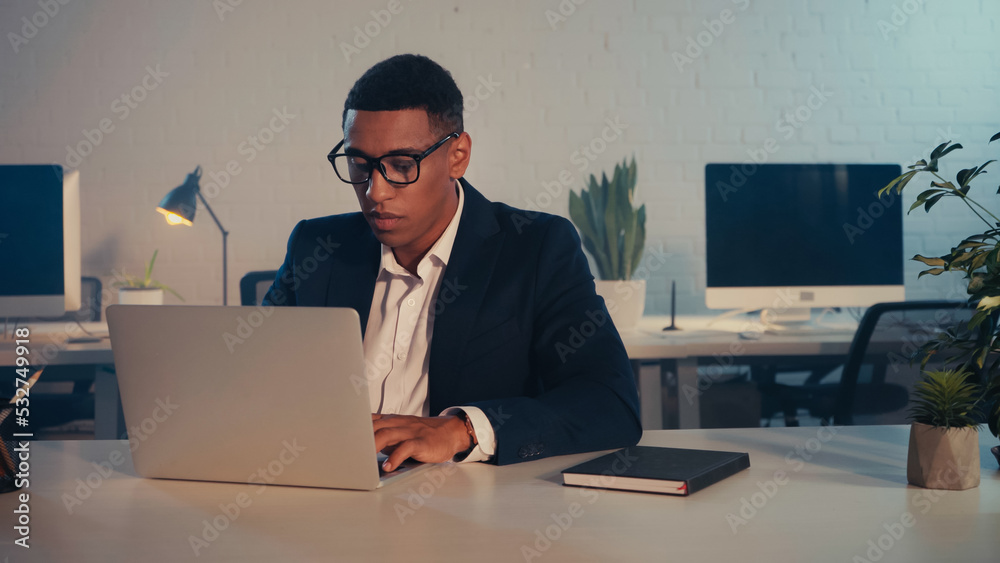 African american businessman in eyeglasses using laptop in office in evening. African american businessman in eyeglasses using laptop in office in evening.