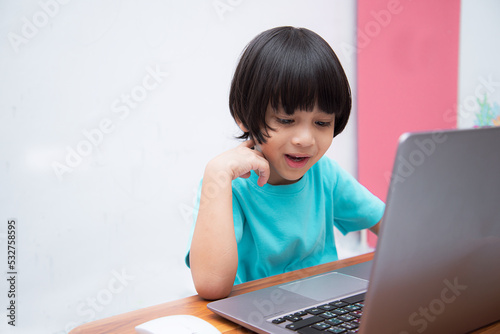 Cute smart Asian boy is happily using his laptop to study online by himself at home. Online learning concept.Self-Study.