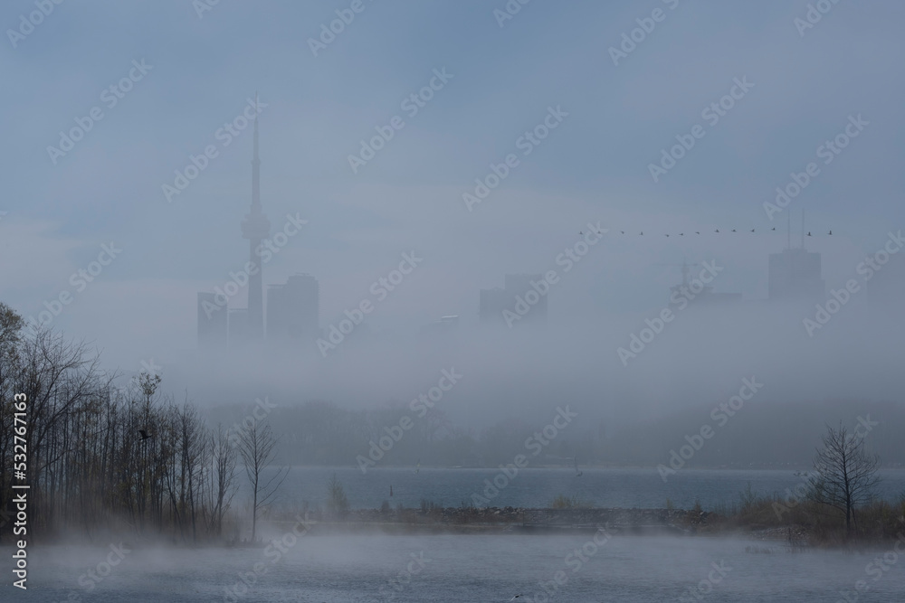 Fototapeta premium Fog and mist over lake Ontario water surface covers Toronto city skyline, heavy clouds on the sky. Weather conditions, advisory, alert concept. Space for copy.