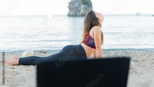 Young beautiful woman practicing yoga, lying in Cobra pose, doing Bhujangasana exercise, attractive girl in leggings and bra working out at the beach following an online yoga class via laptop.