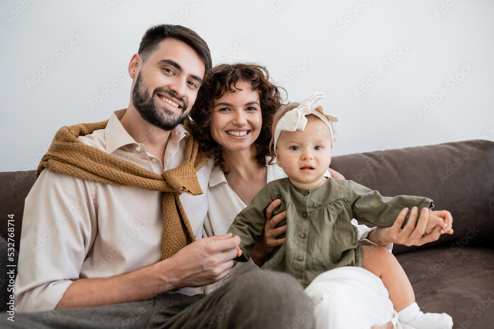 positive parents and infant girl looking at camera while sitting on sofa in living room.