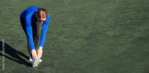 Women and sport. Girl in sportswear does exercises: bends and stretches on the grass at the stadium on a sunny day. Middle aged sportswoman dressed in sportsclothes exercising outdoors. Banner