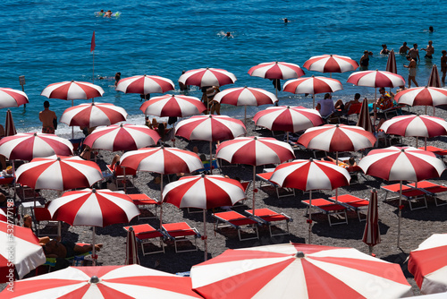 Fototapeta Naklejka Na Ścianę i Meble -  Amalfi beach on famous Amalfi Coast Italy. Rows of red and white parasols or beach umbrellas and tourists relaxing in the sun or bathing in the turquoise sea. Popular holiday destination near Naples.