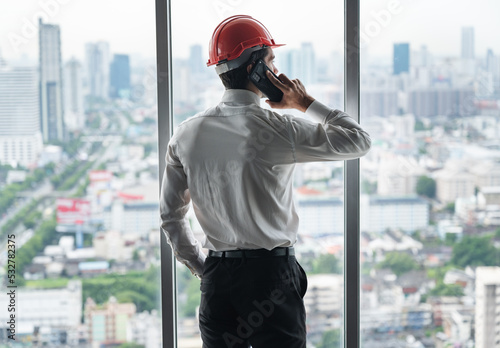 Handsome engineer with smart phone and hardhat looking to the city outside window 