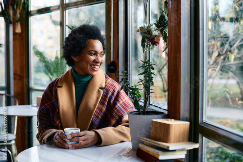 Happy black woman looks through window while enjoying in cup of warm tea in cafe during winter season.