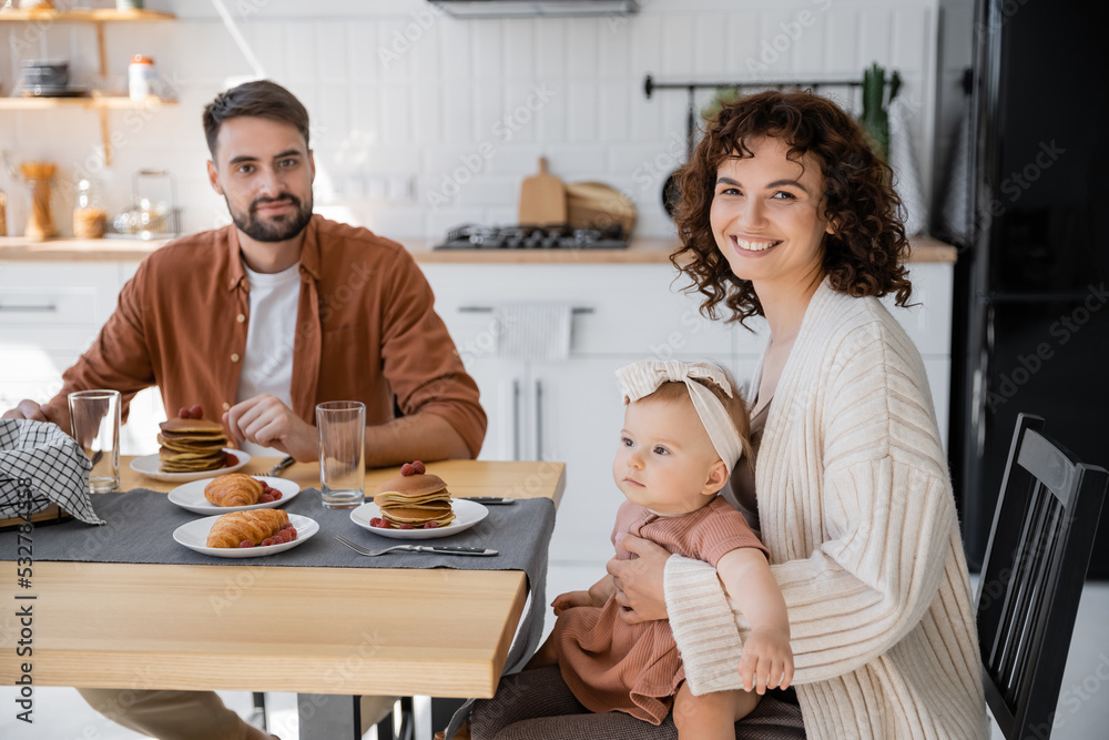 cheerful mother holding infant daughter and sitting at table with husband during breakfast. cheerful mother holding infant daughter and sitting at table with husband during breakfast.