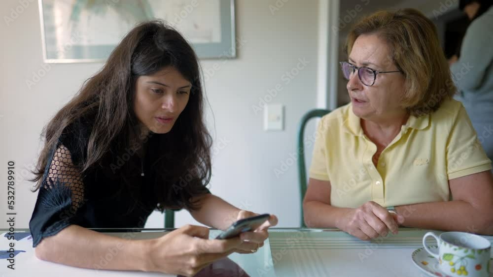 Two people looking at cellphone screen. Older woman putting reading glasses. Person browsing internet online sharing screen with senior mother Two people looking at cellphone screen. Older woman putting reading glasses. Person browsing internet online sharing screen with senior mother