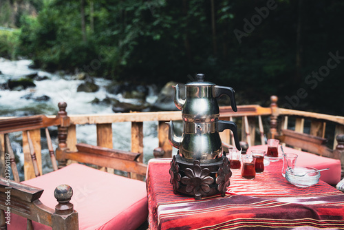 traditional Turkish tea chrome kettle and glasses served on a restaurant table with chairs near a river in an outdoor setting