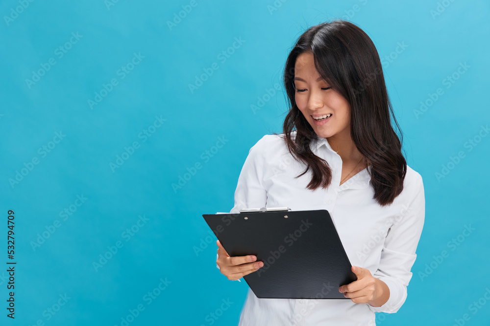 Happy cute Asian businesswoman in classic office dress code holds folder tablet read candidate's resume posing isolated on over blue studio background. Cool business offer. Job interview concept Happy cute Asian businesswoman in classic office dress code holds folder tablet read candidate's resume posing isolated on over blue studio background. Cool business offer. Job interview concept