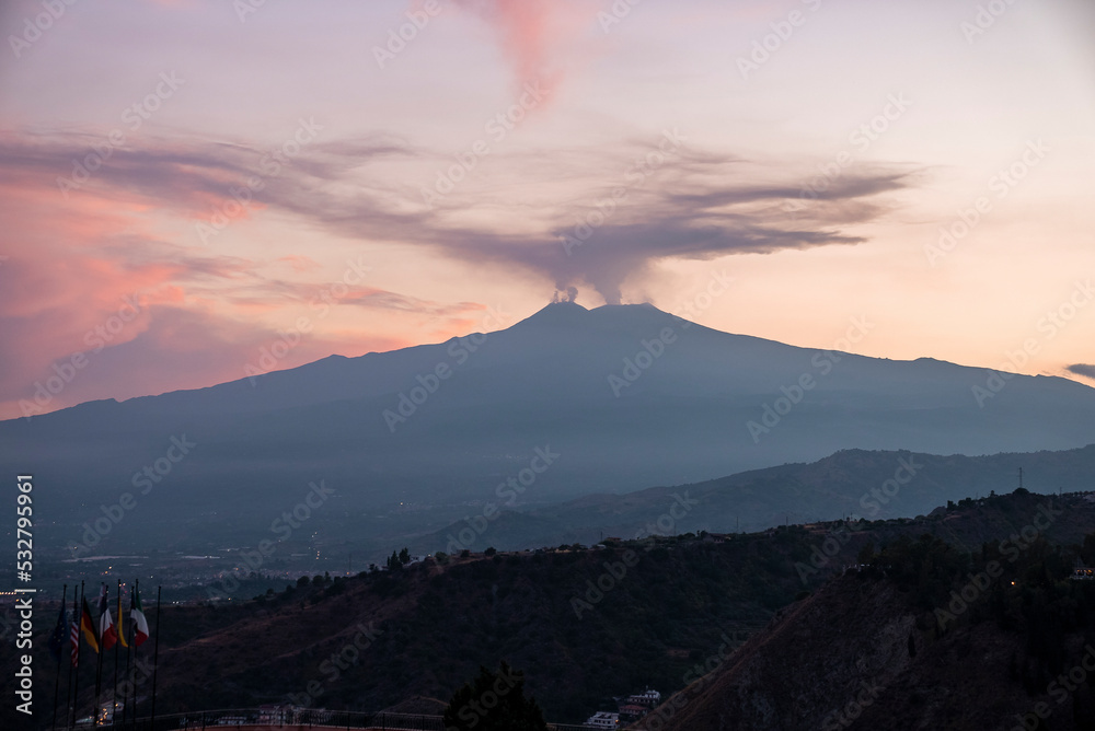 Fototapeta premium Smoke emitting from Mount Etna with orange sky in background. Beautiful tourist attraction with luxurious Elios hotel in foreground. Scenic view of dramatic landscape during sunset.