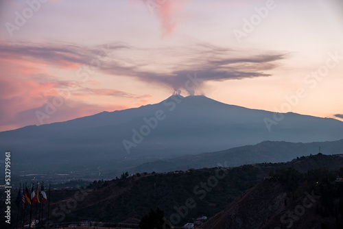 Smoke emitting from Mount Etna with orange sky in background. Beautiful tourist attraction with luxurious Elios hotel in foreground. Scenic view of dramatic landscape during sunset. photo