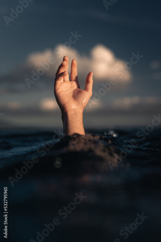 Front of man's hand reaching out of water of blue ocean at sunset with puffy white clouds in background, conceptual image with guy's hand just above wave symbolizing sinking © Lucas
