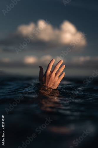 Back of man's hand reaching out of water of blue ocean at sunset with puffy white clouds in background, conceptual image with guy's hand just above wave symbolizing sinking © Lucas