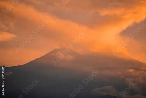 Beautiful volcanic Mount Etna covered with smoke during sunset. Orange clouds wrapped around peak at twilight. Breathtaking scenery of travel place with atmospheric sky in background.