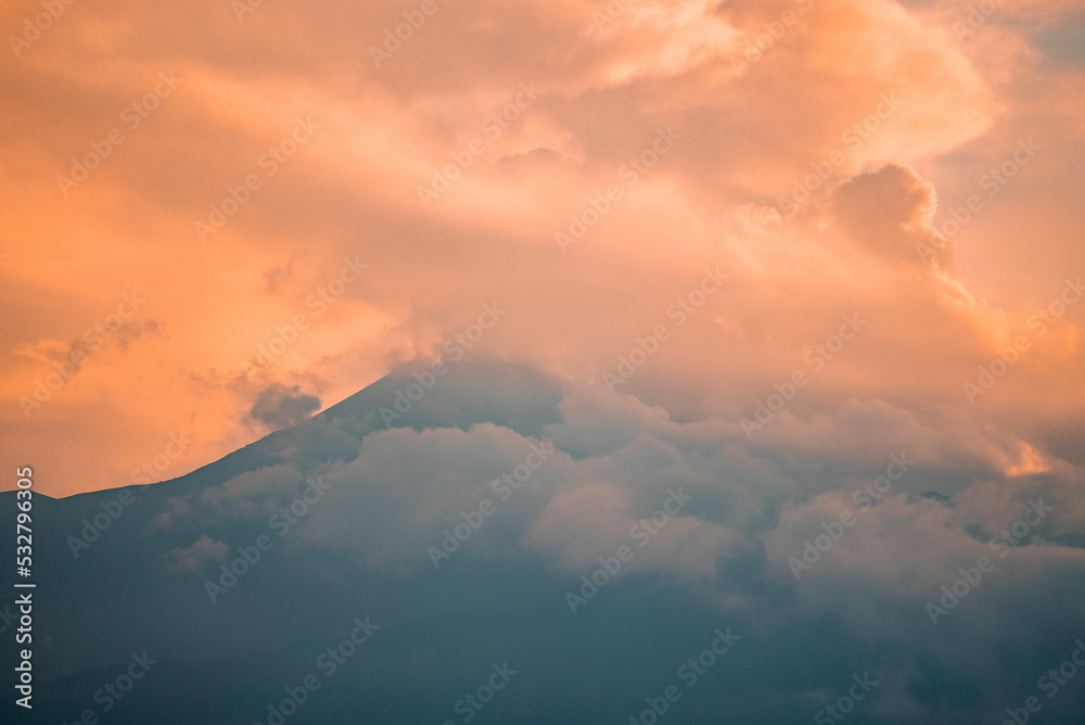 Scenic view of Mount Etna covered with smoke. Vibrant clouds wrapped around peak at twilight. Picturesque scenery of famous tourist attraction with orange sky in background during sunset.