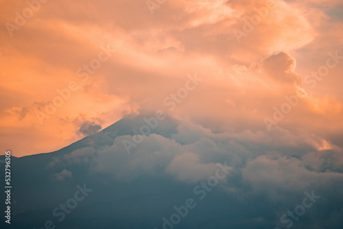 Scenic view of Mount Etna covered with smoke. Vibrant clouds wrapped around peak at twilight. Picturesque scenery of famous tourist attraction with orange sky in background during sunset.