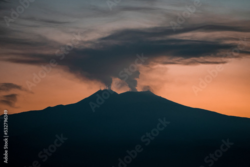 View of smoke emitting from famous Mount Etna with orange sky in background. Atmospheric clouds covering mountain peak. Picturesque scenery of famous tourist attraction during sunset.