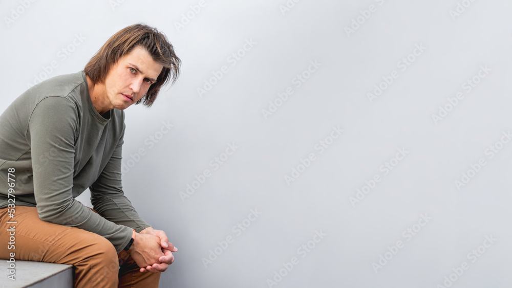 Millennial man with long hair and tense look sits near grey wall. Serious man in earth toned casual clothes is staring in camera. Copy space. Millennial man with long hair and tense look sits near grey wall. Serious man in earth toned casual clothes is staring in camera. Copy space.