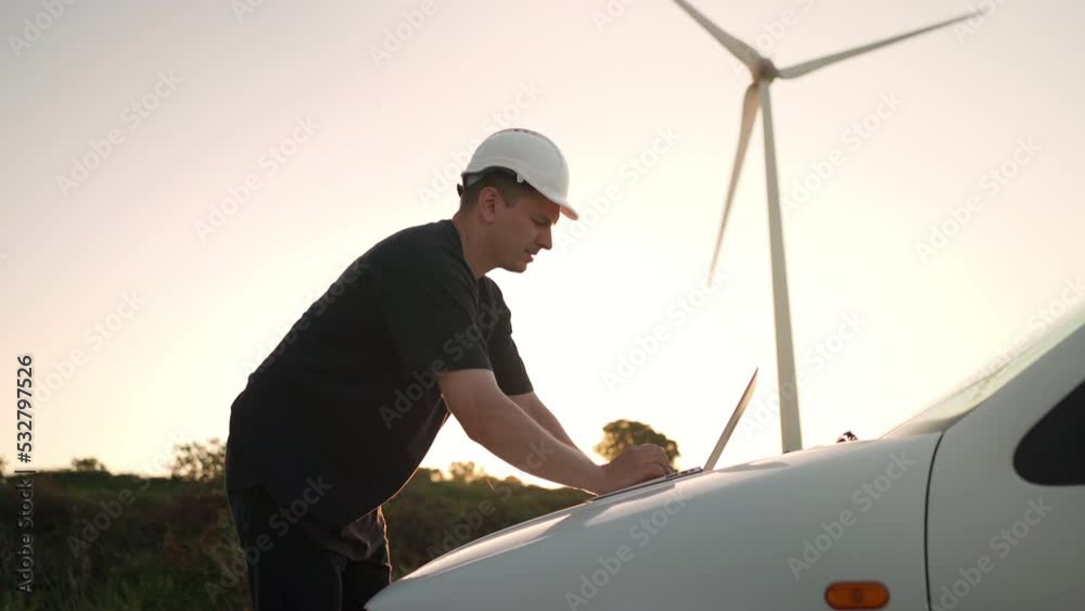 Technician engineer working at wind turbine using laptop computer worker or operator checks wind turbine farm does maintenance to power generator station lean on car wind turbine and sunset background