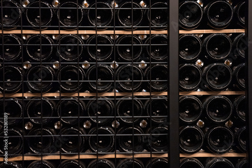 Glass bottles of wine arranged on wooden shelves for aging. Liquor stored together in wine making cellar. Concept of grape juice fermentation for tasty beverage.