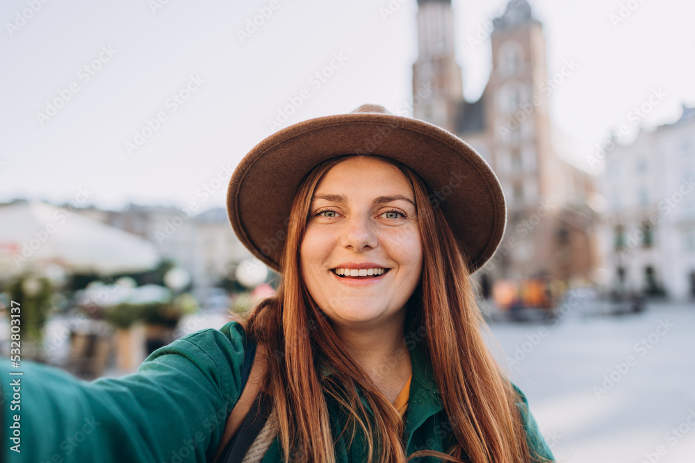 Young woman tourist in hat making selfie photo in front of the famous St. Mary's Basilica on the Market square in Krakow, Poland. Traveling Europe in autumn.