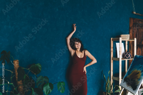 Cheerful woman with raised hand standing near blue wall