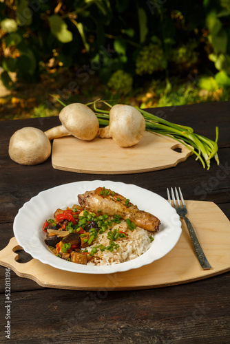Rice with stewed vegetables in sauce and chicken leg in a white plate on a wooden table next to champignons and green onions on the board.