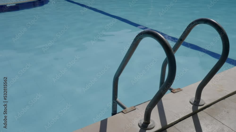 Poolside with stairs and handrail against blue water in tiled swimming pool in summer. Static footage of ladder leading into refreshing pool at hotel on a sunny day. Hotel facilities at resort. Poolside with stairs and handrail against blue water in tiled swimming pool in summer. Static footage of ladder leading into refreshing pool at hotel on a sunny day. Hotel facilities at resort.