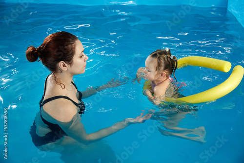 Preschool girllearning to swim in pool with foam noodle with young trainer photo