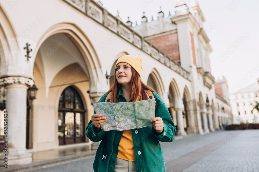 Attractive young female tourist is exploring new city. Redhead girl in hat holding a paper map on Market Square in Krakow. Traveling Europe in autumn.