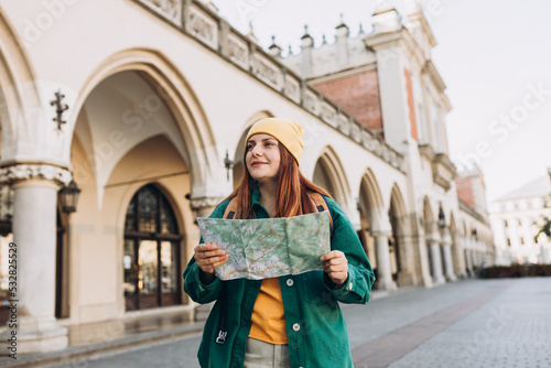 Obraz na płótnie Attractive young female tourist is exploring new city. Redhead girl in hat holding a paper map on Market Square in Krakow. Traveling Europe in autumn. w salonie
