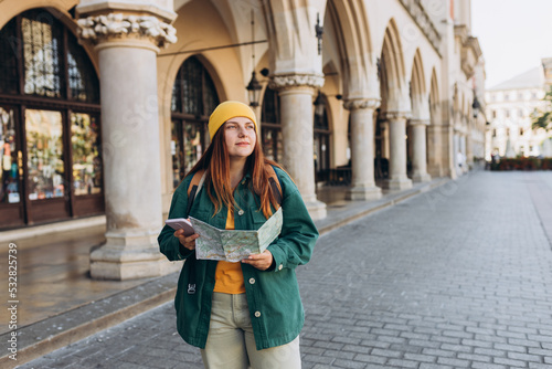 Attractive young female tourist is exploring new city. Redhead girl in hat holding a paper map and smartphone on Market Square in Krakow. Traveling Europe in autumn. Active lifestyle concept © mdyn
