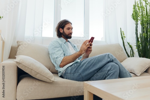 A man with a beard sits on the couch during the day and smiles at home and looks at his phone relaxing on his day off, life online on his phone texting with friends