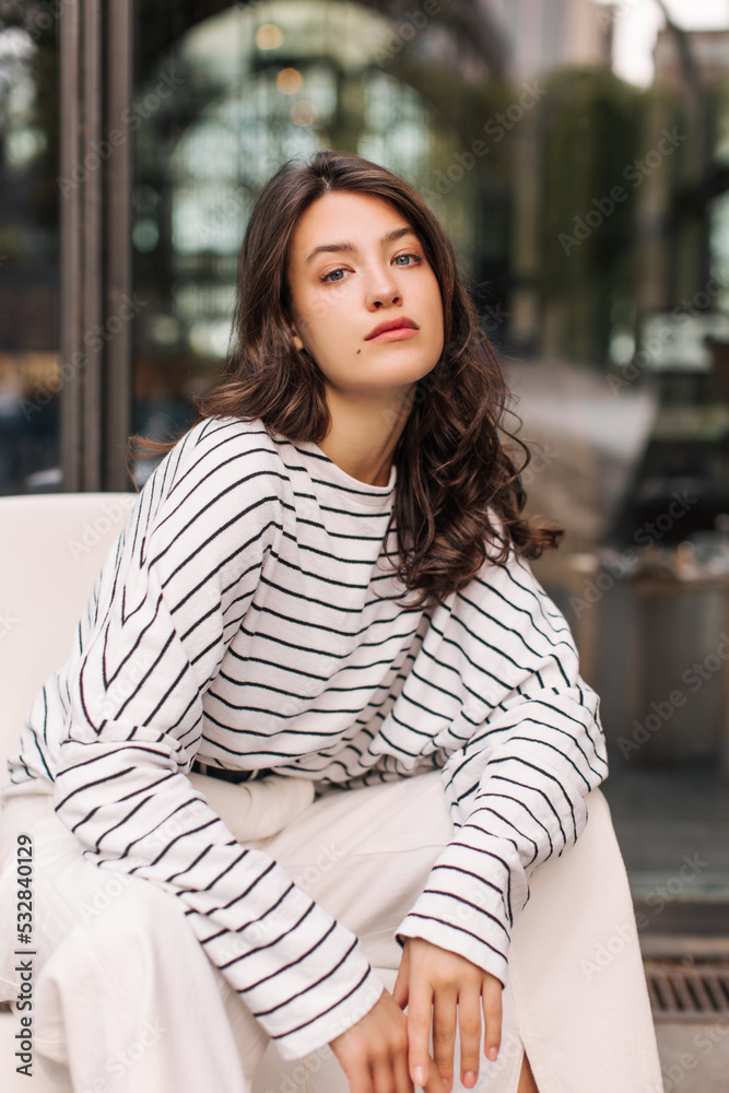 Beautiful young european girl looks at camera spending time sitting near city building. Brunette woman with wavy hair wears casual clothes. Beauty and youth concept